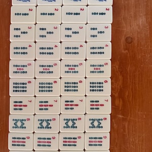 K&ouml;nnte beinhalten: Eine Anordnung wei&szlig;er Mahjong-Steine auf einer Holzoberfl&auml;che. Jede Kachel zeigt eine Zahl von 1 bis 9 sowie verschiedene blaue und rote Symbole und Illustrationen. Die rechteckigen Kacheln haben abgerundete Ecken.