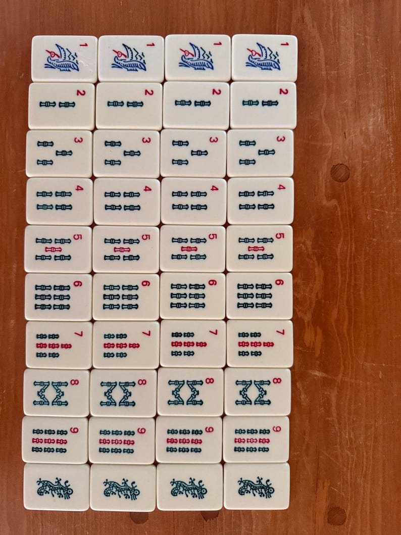 K&ouml;nnte beinhalten: Eine Anordnung wei&szlig;er Mahjong-Steine auf einer Holzoberfl&auml;che. Jede Kachel zeigt eine Zahl von 1 bis 9 sowie verschiedene blaue und rote Symbole und Illustrationen. Die rechteckigen Kacheln haben abgerundete Ecken.