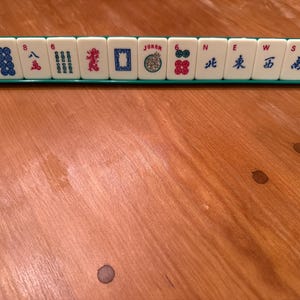 K&ouml;nnte beinhalten: Ein Satz Mahjong-Steine, der auf einem gr&uuml;nen Gestell angeordnet ist. Die wei&szlig;en Steine weisen verschiedene Symbole, Zahlen und chinesische Schriftzeichen in Blau, Rot und Gr&uuml;n auf. Ein Stein tr&auml;gt die Aufschrift "JOKER".