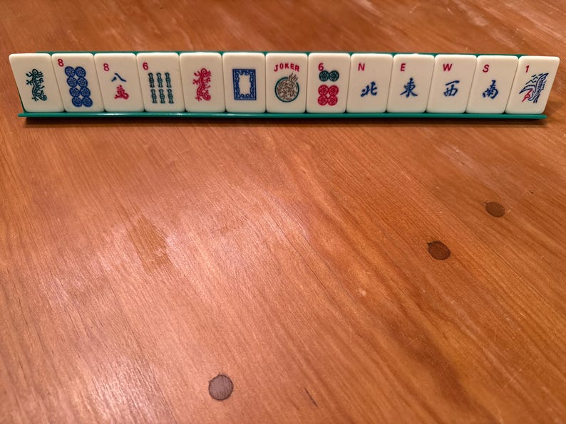 K&ouml;nnte beinhalten: Ein Satz Mahjong-Steine, der auf einem gr&uuml;nen Gestell angeordnet ist. Die wei&szlig;en Steine weisen verschiedene Symbole, Zahlen und chinesische Schriftzeichen in Blau, Rot und Gr&uuml;n auf. Ein Stein tr&auml;gt die Aufschrift "JOKER".