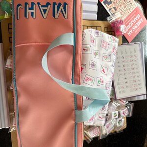 May include: A pink Mahjong set with a matching carrying case. The case has the word "MAHJ" in blue block letters. The set includes tiles, dice, and a printed guide. The case has a light blue handle and trim.