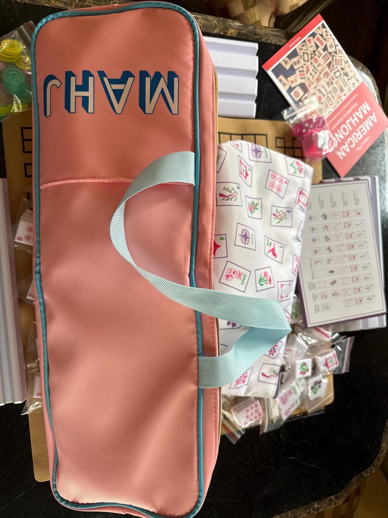 May include: A pink Mahjong set with a matching carrying case. The case has the word "MAHJ" in blue block letters. The set includes tiles, dice, and a printed guide. The case has a light blue handle and trim.