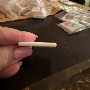 May include: A small, rectangular, white object is held between fingers. The object appears to be a small tile or a similar craft supply. Other similar objects are visible in the background, packaged in clear plastic bags.