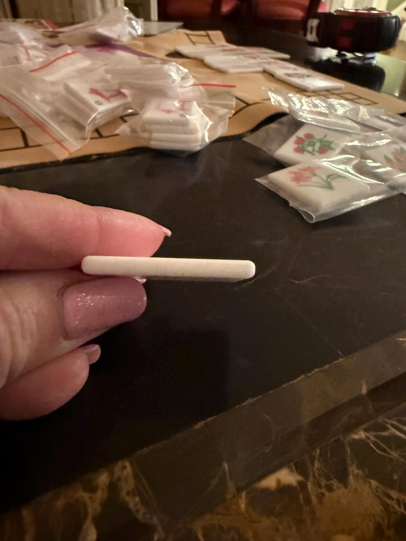 May include: A small, rectangular, white object is held between fingers. The object appears to be a small tile or a similar craft supply. Other similar objects are visible in the background, packaged in clear plastic bags.