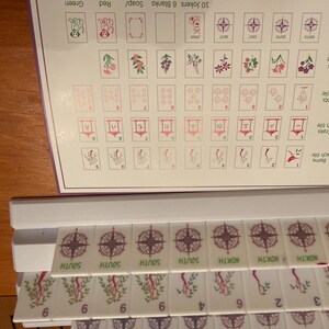 May include: A close-up of a Mahjong tile set. The tiles are white with various symbols and numbers. The set includes tiles with compass rose designs, bamboo, and other suits. A reference sheet is visible.
