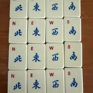K&ouml;nnte beinhalten: Zw&ouml;lf cremefarbene Mahjong-Steine, angeordnet in einem Raster. Jeder Stein zeigt einen Buchstaben (N, E, W oder S) und ein blaues chinesisches Schriftzeichen. Die Steine sind rechteckig mit abgerundeten Ecken, vor einem warmbraunen Hintergrund.