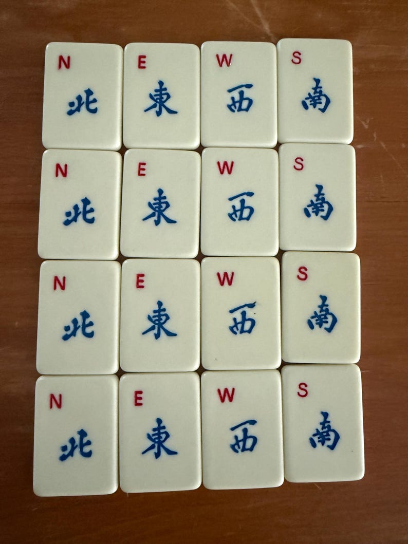 K&ouml;nnte beinhalten: Zw&ouml;lf cremefarbene Mahjong-Steine, angeordnet in einem Raster. Jeder Stein zeigt einen Buchstaben (N, E, W oder S) und ein blaues chinesisches Schriftzeichen. Die Steine sind rechteckig mit abgerundeten Ecken, vor einem warmbraunen Hintergrund.