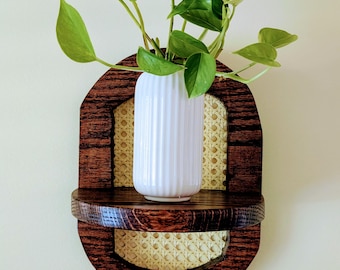 Puede incluir: Un estante de pared para plantas con un marco de madera oscura y un respaldo de mimbre tejido. Un jarrón de cerámica blanco y acanalado contiene una planta verde y frondosa. El estante tiene un diseño curvo.