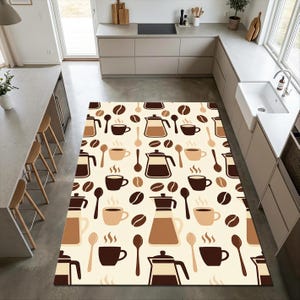 Op de afbeelding: Een rechthoekig vloerkleed met een koffiethema, met een crèmekleurige achtergrond. Het patroon bevat koffiebonen, kopjes, lepels en koffiepotten in bruine en beige tinten. Het kleed ligt in een keuken.