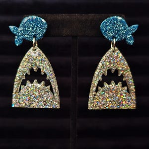 May include: A pair of glittery shark earrings. The earrings feature a blue glitter fish-shaped top and a silver glitter shark jaw pendant. The shark jaw pendant has a jagged tooth design. The earrings are displayed on a black stand.