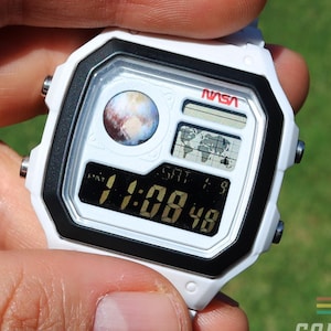 May include: White digital watch with a black bezel and a white band. The watch face displays the NASA logo, a planet graphic, and the time 11:08:48. The watch is held in a hand against a green background. The text "COLORCHRONOS" is visible.