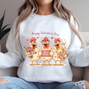 May include: White sweatshirt with a Valentine's Day graphic featuring three cartoon chickens. The chickens hold a bouquet of flowers, a sign that says "Be Mine", and a teddy bear. The text "Happy Valentine's Day" is above the chickens.