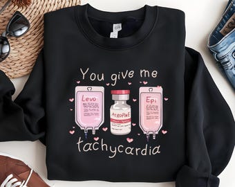 May include: Black sweatshirt with the phrase "You give me tachycardia" and illustrations of medical supplies. The design includes pink IV bags labeled "Levo" and "Epi", and a vial of "Atropine". Small pink hearts are scattered around the text and images.