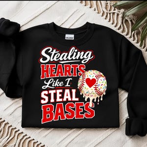 May include: Black sweatshirt with the text "Stealing HEARTS Like I STEAL BASES" in white and red lettering. A baseball with a heart and sprinkles is on the right side. The sweatshirt is made of a soft material.