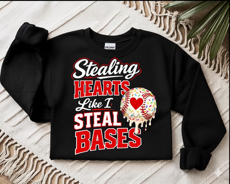 May include: Black sweatshirt with the text "Stealing HEARTS Like I STEAL BASES" in white and red lettering. A baseball with a heart and sprinkles is on the right side. The sweatshirt is made of a soft material.