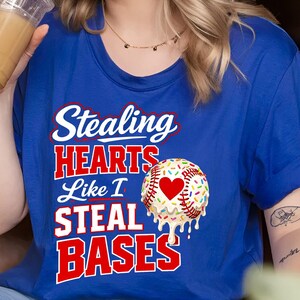 May include: Royal blue t-shirt with the text "Stealing HEARTS Like I STEAL BASES" in white and red. A baseball with a heart and sprinkles is on the right side of the shirt. The shirt is a baseball-themed design.