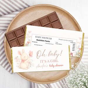 Puede incluir: Una barra de chocolate y un envoltorio de barra de caramelo con temática de baby shower con el texto "Oh baby! It's a girl". El envoltorio presenta un conejito sosteniendo una mariposa, con detalles florales.