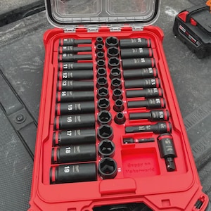 May include: A red Milwaukee PACKOUT organizer case with a clear lid containing a set of black impact sockets. The interior features a red molded tray holding various socket sizes, with the Milwaukee logo visible on the lid and tools.