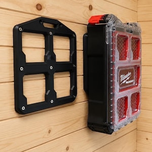 Könnte beinhalten: Eine schwarze Kunststoff-Montageplatte mit vier quadratischen Öffnungen und ein Milwaukee Packout-Organizer-Koffer mit transparentem Deckel, roten Inneneinteilungen und rotem Verschluss, montiert an einer hellen Holzwand.