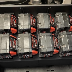 Könnte beinhalten: Acht schwarz-rote Milwaukee M18 5.0 Lithium-Ionen-Akkus für Elektrowerkzeuge, angeordnet in zwei Reihen zu je vier Stück in einem schwarzen Aufbewahrungskoffer. Jeder Akku verfügt über einen roten Entriegelungsknopf, eine Ladezustandsanzeige sowie den Schriftzug M18 und 5.0.