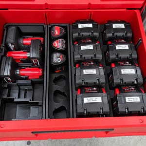 Op de afbeelding: Een rode gereedschapslade met zwarte kunststof organizers. Het linkergedeelte bevat twee rood-zwarte elektrische gereedschappen. Het middengedeelte bevat drie kleine ronde voorwerpen. Rechts bevinden zich acht zwarte accupacks met witte labels.