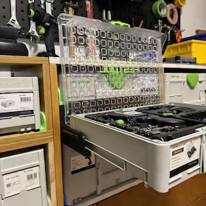 May include: A clear plastic tool organizer with a grid pattern is pulled out of a gray storage container. The container is part of a modular storage system. Tools and equipment are visible in the background, mounted on a black pegboard.