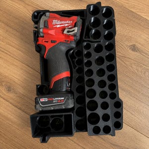 Könnte beinhalten: Ein roter und schwarzer Milwaukee Fuel Akku-Schlagschrauber mit einem M12 Redlithium XC 3.0 Akku, platziert in einer schwarzen Kunststoff-Aufbewahrungsschale mit mehreren kreisförmigen Schlitzen und numerischen Beschriftungen zur Werkzeugorganisation.