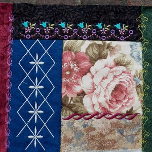 Può includere: Un blocco di patchwork per trapunta con un motivo floreale al centro, circondato da un tessuto marrone scuro con fiori ricamati, un tessuto blu con rombi ricamati bianchi, un tessuto bordeaux con cuciture rosa e un tessuto verde con cuciture gialle.