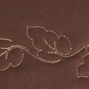 May include: A brown background with a delicate embroidered design of three leaves connected by a curving stem. The leaves and stem are stitched with a light gold thread, creating a subtle contrast against the darker backdrop.