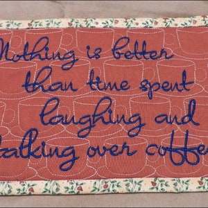 以下が含まれることがあります： 赤い背景に白いコーヒーカップの輪郭が描かれた、長方形のキルトプレースマット。「Nothing is better than time spent laughing and talking over coffee」というテキストが青色で刺繍されています。縁には花柄があります。