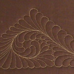 Può includere: Un tessuto marrone con un disegno ricamato color oro. Il disegno è un motivo stilizzato a piuma o foglia, disposto a forma triangolare. Il ricamo è dettagliato e intricato, con un effetto a linea tratteggiata.