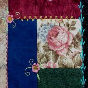 Può includere: Un blocco di trapunta patchwork con un motivo floreale al centro. Il blocco è circondato da un bordo di tessuto blu, verde e rosso. Il tessuto è cucito insieme con una cucitura decorativa.