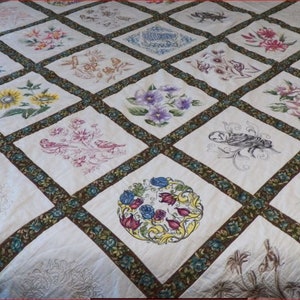 Op de afbeelding: Een wit quilt met een bruine rand en verschillende geborduurde bloemen- en vogelmotieven. Het quilt is gemaakt van 16 vierkanten, elk met een ander motief.