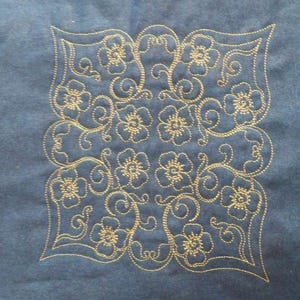 May include: A square, embroidered design featuring a floral pattern in gold thread on a blue fabric background. The design includes flowers and swirling vines, creating a symmetrical and decorative motif. The embroidery is detailed and intricate.