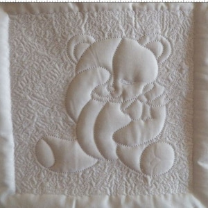 May include: A white quilted fabric square with a white embroidered design of a bear holding a heart. The bear is in the centre of the square and the heart is in its paws.