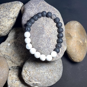 May include: A beaded bracelet featuring alternating black and white lava stone beads. The bracelet has a silver-colored bead and is displayed on a background of gray rocks. The bracelet is a stylish accessory.