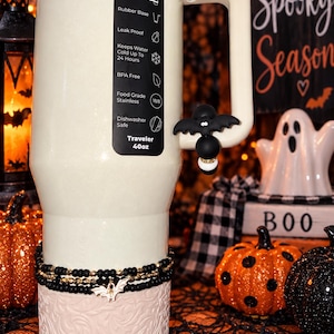 May include: A 40oz cream-colored stainless steel tumbler with a handle, featuring a black bat-shaped charm on the handle and a stack of black and gold beaded bracelets with a small bat charm around the base. The tumbler has a black label listing features.