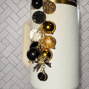 May include: A white tumbler with a gold rim and a decorative charm. The charm features black, gold, and clear beads, a gold bow, and a small black pendant with the letter "J". The charm is attached to the tumbler's handle.