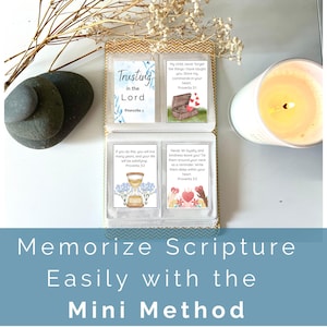 Op de afbeelding: Een klein, open boek met vier kaarten met bijbelverzen en illustraties. De kaarten zitten in een doorzichtige plastic hoes. De tekst "Memorize Scripture Easily with the Mini Method" staat onderaan. Een brandende kaars, stenen en droogbloemen staan ook in de afbeelding.