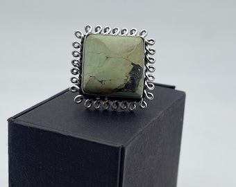 May include: A square ring featuring a pale green stone with dark brown and black markings, set within a silver-toned, swirled border. The ring is displayed on a black box against a white backdrop.