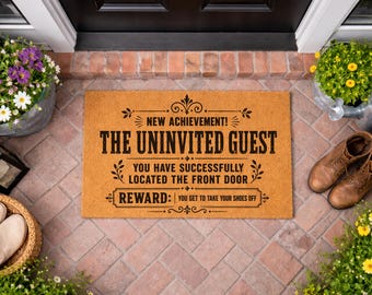 May include: A rectangular tan coir doormat with black text that reads: NEW ACHIEVEMENT! THE UNINVITED GUEST YOU HAVE SUCCESSFULLY LOCATED THE FRONT DOOR REWARD: YOU GET TO TAKE YOUR SHOES OFF. The mat features decorative flourishes and borders.