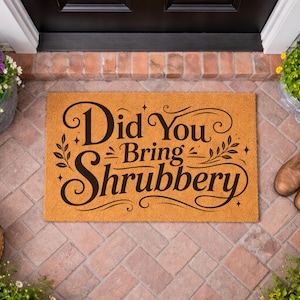 Può includere: Un zerbino rettangolare con la scritta nera "Did You Bring Shrubbery" su sfondo beige. Il tappeto è su un patio di mattoni, circondato da piante, scarpe e una lanterna.