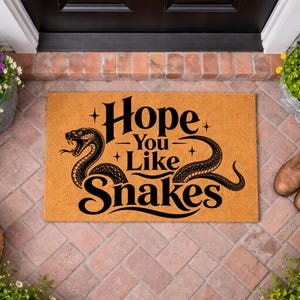 May include: A rectangular tan coir doormat featuring a black graphic of a coiled snake on the left and the text "Hope You Like Snakes" in a stylised font. The design includes small four-pointed star accents surrounding the bold black lettering.