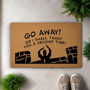 Peut inclure: Un paillasson rectangulaire avec le texte "GO AWAY! OR I SHALL TAUNT YOU A SECOND TIME!" en noir. Le tapis présente une silhouette noire d'une personne pointant et un motif de mur de briques sur un fond beige.