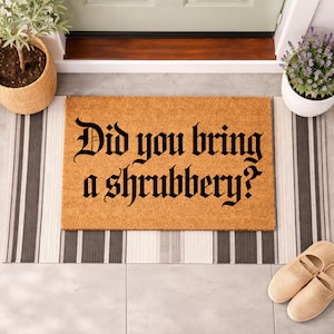 May include: A rectangular coir doormat with the question "Did you bring a shrubbery?" in black gothic font. The mat rests on a striped rug, with potted plants and slippers visible, creating a welcoming entrance.