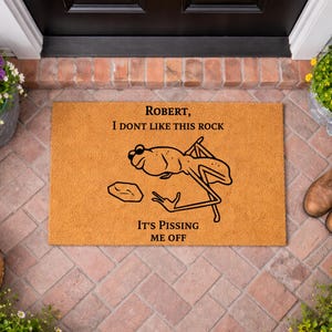 May include: A rectangular tan coir doormat featuring a black line drawing of a frog wearing sunglasses next to a rock. The text reads: ROBERT, I DONT LIKE THIS ROCK IT'S PISSING ME OFF. The mat has a coarse, natural fiber texture.