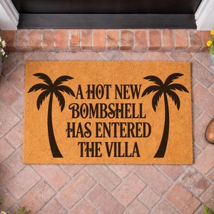 May include: A rectangular tan coir doormat featuring two black palm tree silhouettes on the sides and the black text A HOT NEW BOMBSHELL HAS ENTERED THE VILLA centered between the trees in a decorative serif font style.