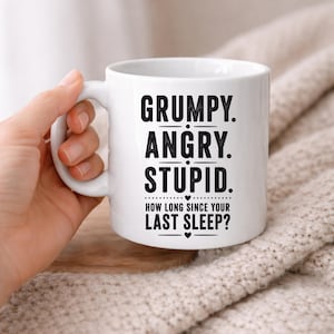 May include: White ceramic mug with the words "GRUMPY. ANGRY. STUPID." in bold black letters. Below the words is the text "HOW LONG SINCE YOUR LAST SLEEP?" in black. The mug is held by a hand.