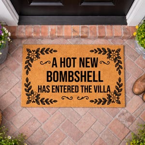 May include: A rectangular tan coir doormat with black text that reads A HOT NEW BOMBSHELL HAS ENTERED THE VILLA. The mat features a decorative black floral and leaf border design in each corner and is centered on a brick porch floor.