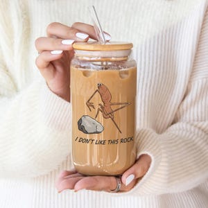 May include: A glass tumbler with a bamboo lid and a clear glass straw, filled with a light brown beverage. The tumbler features a cartoon illustration of a creature and a rock, with the text "I DON'T LIKE THIS ROCK".